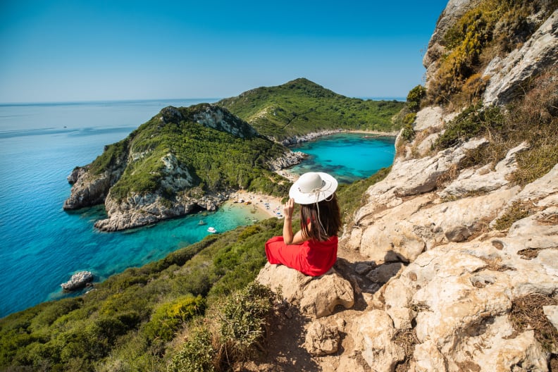 Frau mit Sonnenhut sitzt hoch oben auf einem Felsen und schaut auf Meer © iStock.com/RistoArnaudov